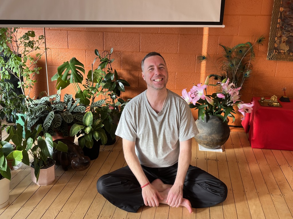 Dejan Kuzmic smiling, sitting cross-legged on the floor of a yoga centre surrounded by plants and flowers